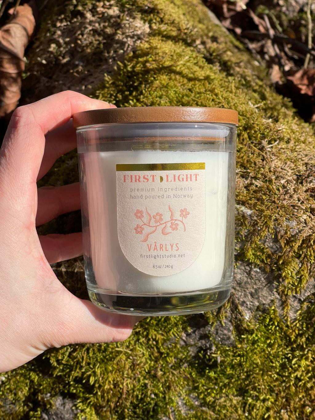 Hand holding a candle labeled 'First Light' by Varly's on a mossy background