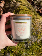 Hand holding a candle labeled 'First Light' by Varly's on a mossy background