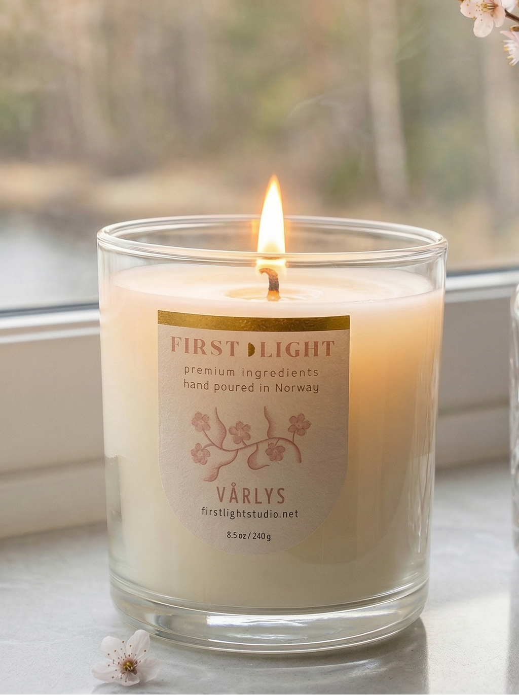 Candle labeled 'First Light' on a windowsill with a vase of flowers.